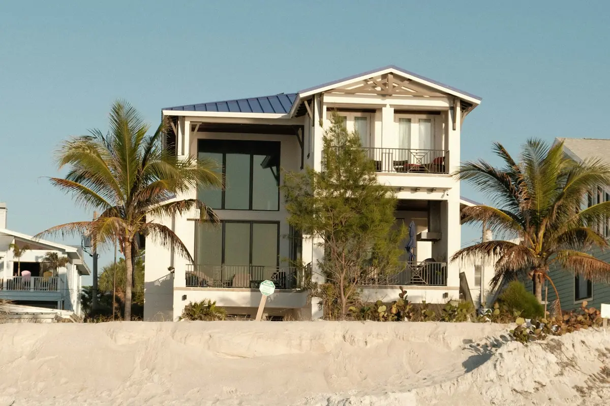 Two-story white beach house with railed balconies and a metal roof, palm trees on either side and a sand dune across the foreground