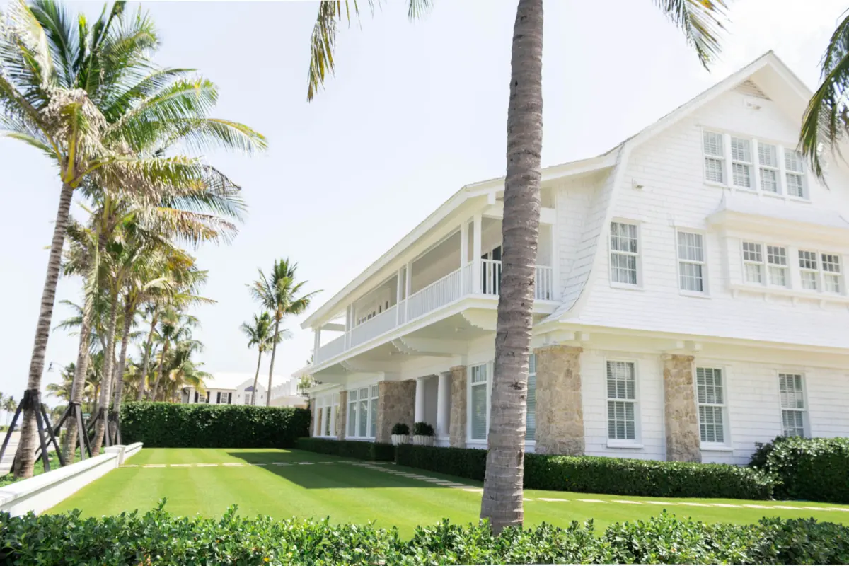 White shingled house with a two-story covered porch on rough stone piers, a clipped hedge and coconut palms across a mown lawn